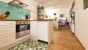 A compact kitchen with white cabinets, a stainless steel oven, and a small refrigerator. There is a wooden countertop with a knife block and a coffee maker. The floor has a patterned rug, and the kitchen leads to a dining area with a table and chairs. - Ferienwohnung Lieblingsplatz (Lueneburg)
