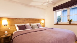 A bedroom with a wooden headboard, a large bed with a purple quilt, bedside lamps, and a window with curtains. - Ferienwohnung Lieblingsplatz (Lueneburg)