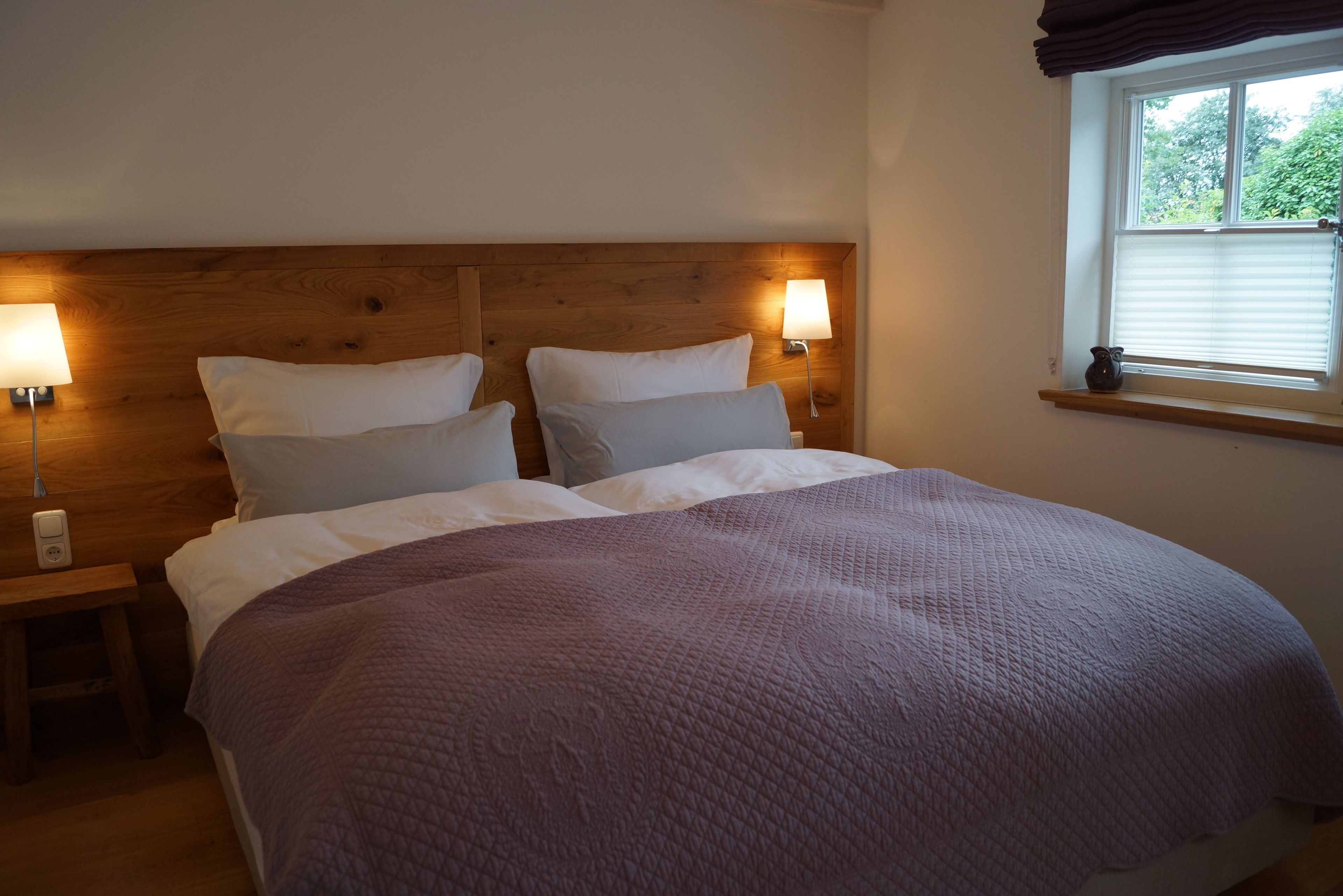 A bedroom with a wooden headboard, two bedside lamps, a window with blinds, and a purple quilted bedspread.