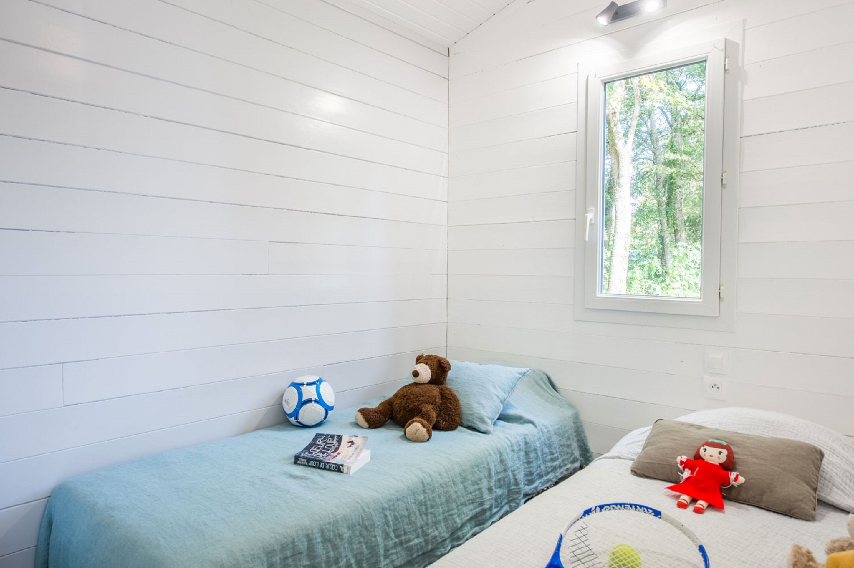 Een kleine kamer met een enkel bed, witte muren en een raam. Op het bed ligt een teddybeer, een boek en een klein speelgoed.