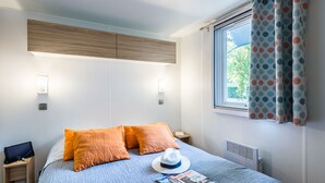 Una cama bien hecha con almohadas anaranjadas, un sombrero blanco y una revista sobre la cama. Se ve una ventana con cortinas y una lámpara fijada a la pared.