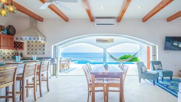 A spacious living area with a view of the ocean, a dining table with chairs, a hammock, and a TV mounted on the wall.
