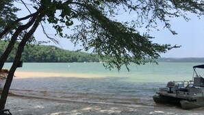A beach with a boat docked on the shore, a sandy area with metal chairs, and a tree providing shade.
