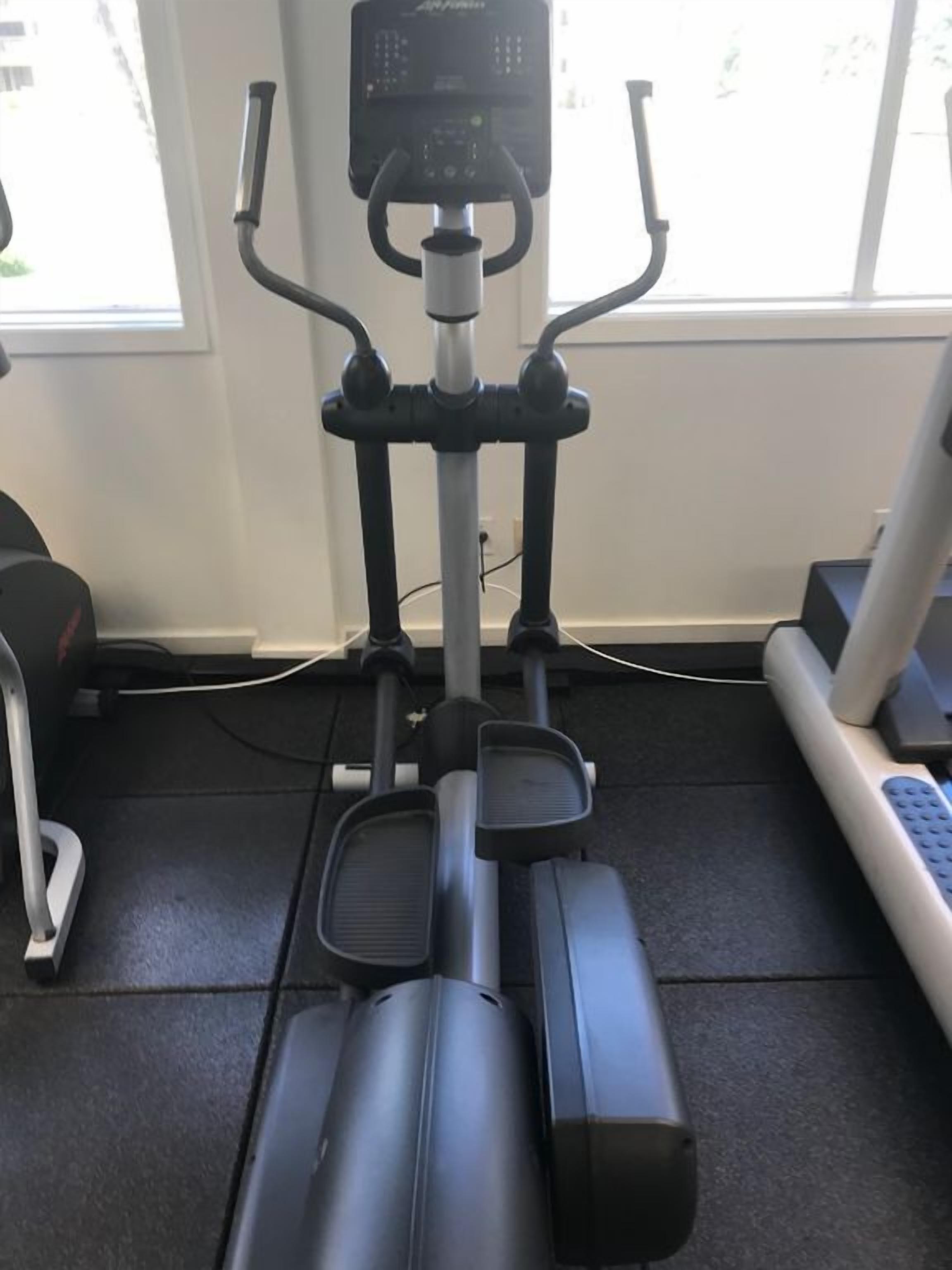 Una bicicleta estática con pantalla digital, ubicada en una habitación con ventana y una caminadora.