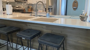 A modern kitchen with white cabinets, stainless steel appliances, and a central island with stools. - Pleasure Island Paradise - closest rental to the beach you will find (Kure Beach)