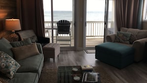 A living room with a view of the ocean through large sliding glass doors, a sofa, a chair, and a coffee table with books. - Pleasure Island Paradise - closest rental to the beach you will find (Kure Beach)