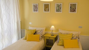 Ein Zimmer mit zwei Betten, jeweils mit gelben Kissen und einem Nachttisch mit einer Lampe. Darüber befinden sich Wandbilder mit floralem Dekor.