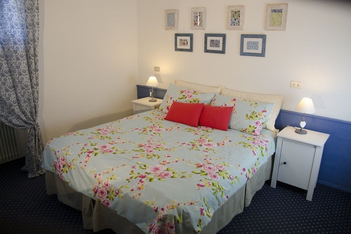 Un dormitorio ordenado con colcha estampada, dos mesitas de noche con lámparas y una pared decorada con cuadros enmarcados.