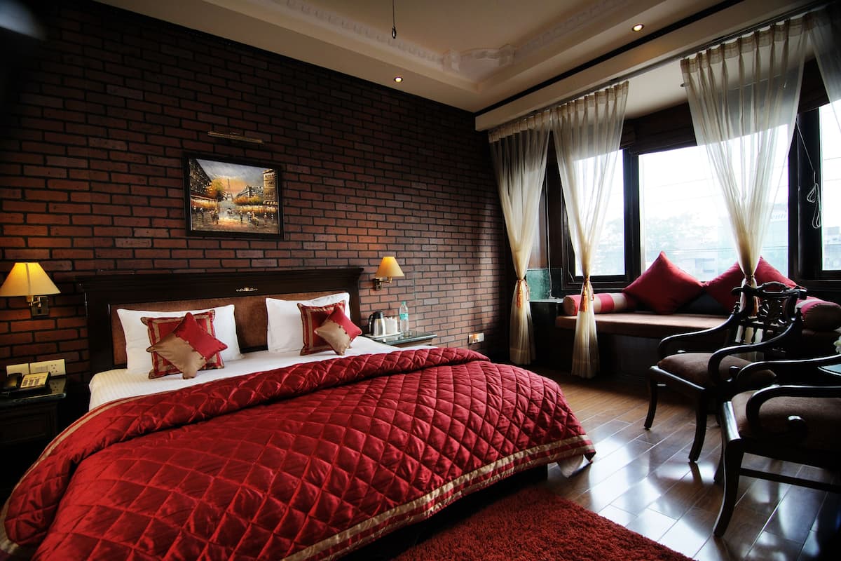 A hotel room with a brick wall, a large bed with red bedding, a nightstand with a lamp, a window with curtains, and a view of a cityscape.