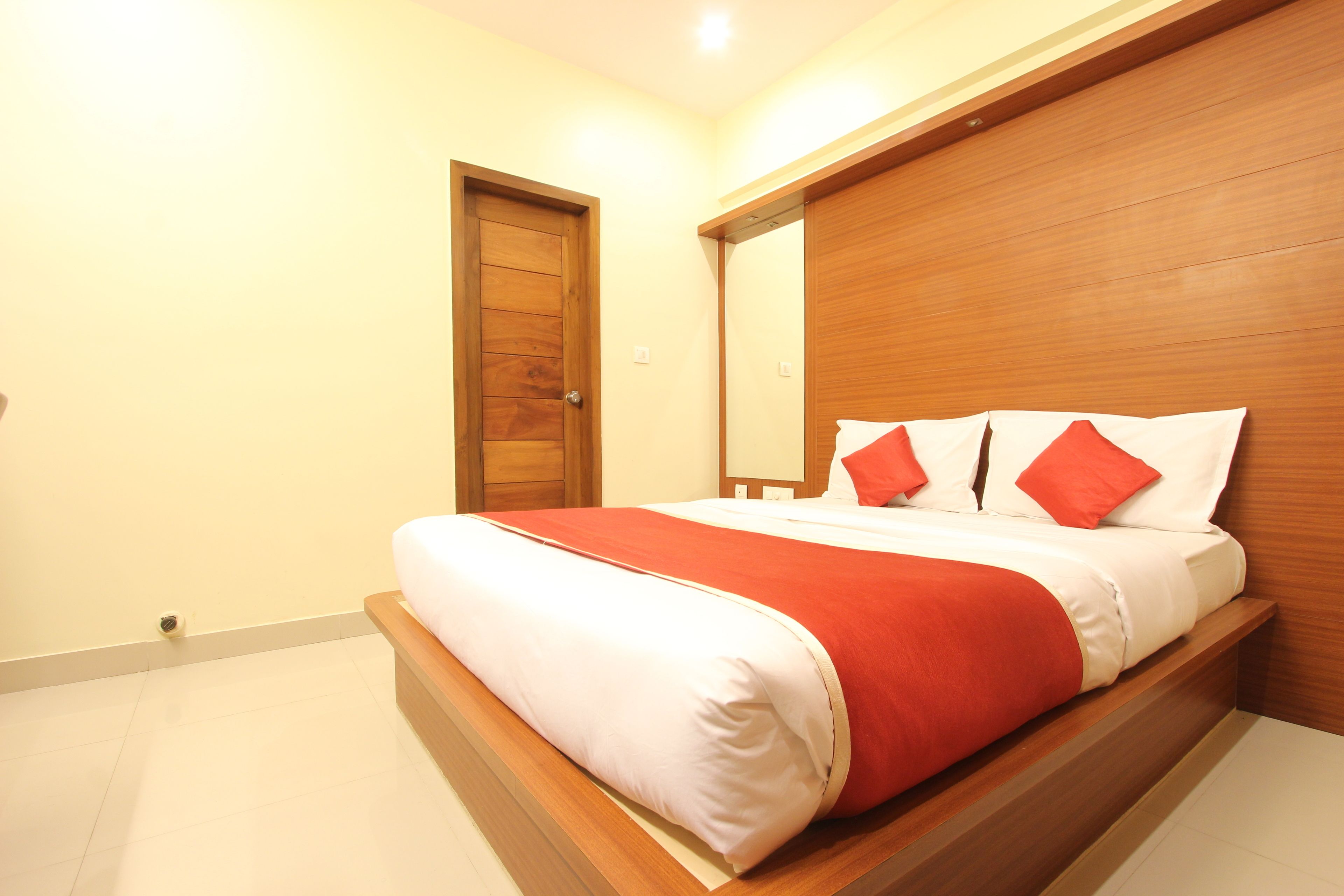 Un dormitorio con cabecera de madera, cama con ropa de cama blanca y roja, y una puerta con cámara de seguridad.