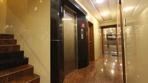 Un ascensor moderno con una señal roja en forma de 'X', una escalinata de mármol y un pasillo limpio y bien iluminado.