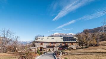 A stone house with a paved driveway, wooden shutters, and a mountain backdrop.