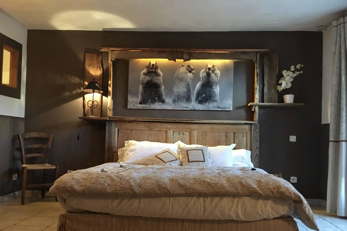 A bedroom with a large bed, a wooden headboard, a wall-mounted picture, a chair, and a shelf with a potted plant.