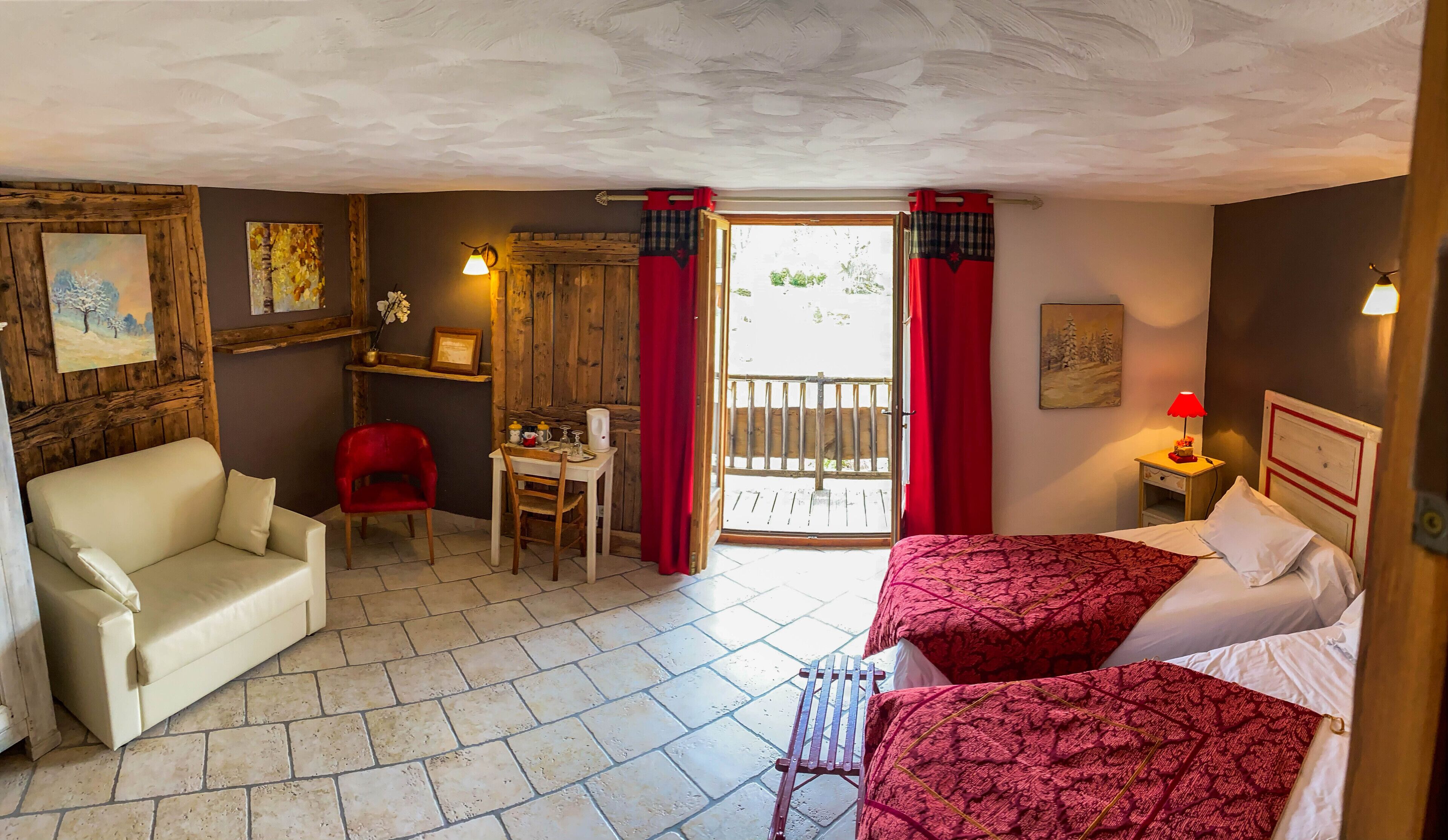 A cozy room with a sofa, a small table with a chair, two beds with red bedding, a wooden shelf with artwork, and a view through a door.