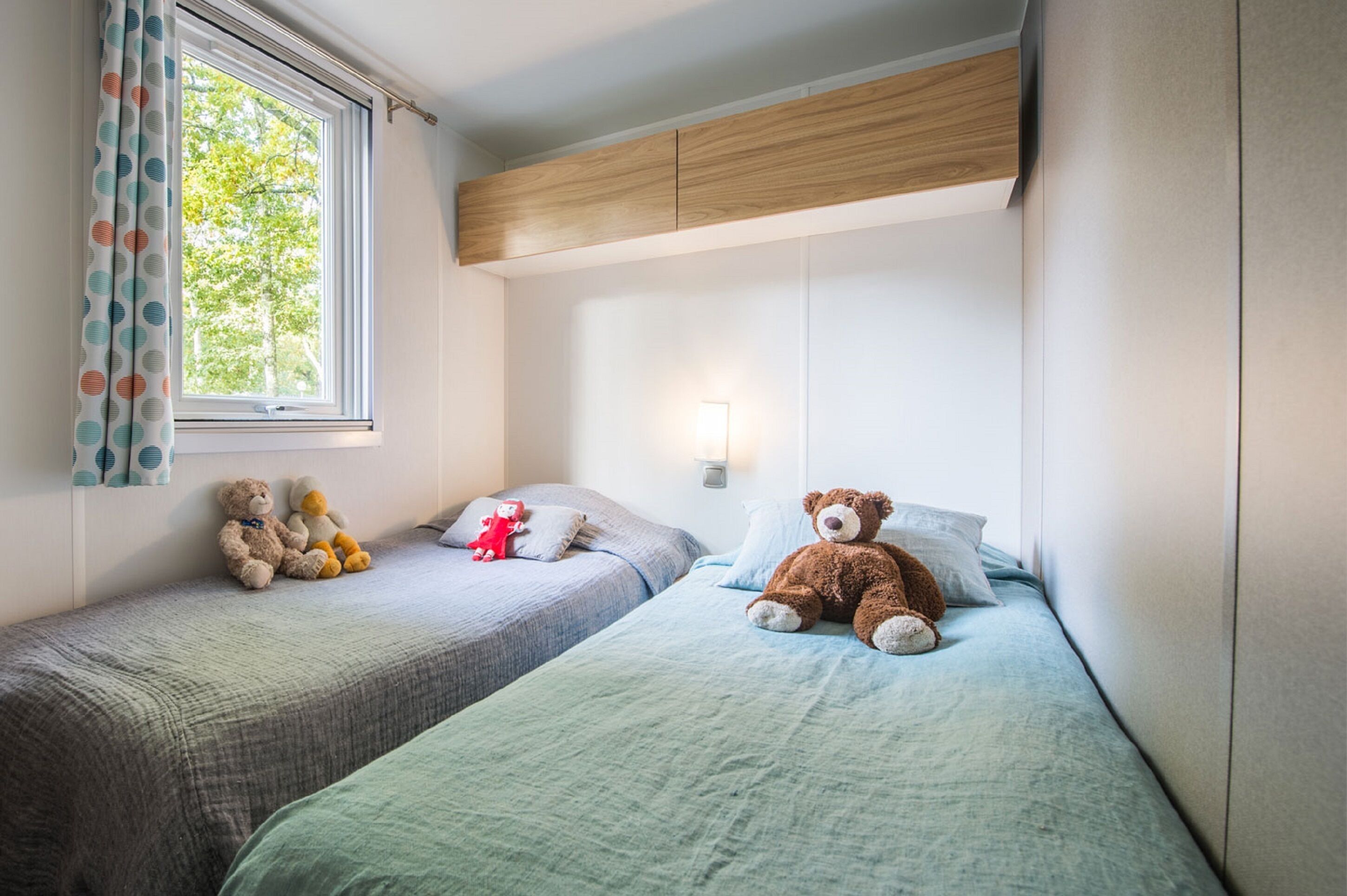 A bedroom with two beds, each with a teddy bear, a window with curtains, and a wall-mounted light.