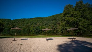 A beach with three thatched umbrellas and a calm lake surrounded by greenery. - Camping le Gibanel (Saint Martial Entraygues)