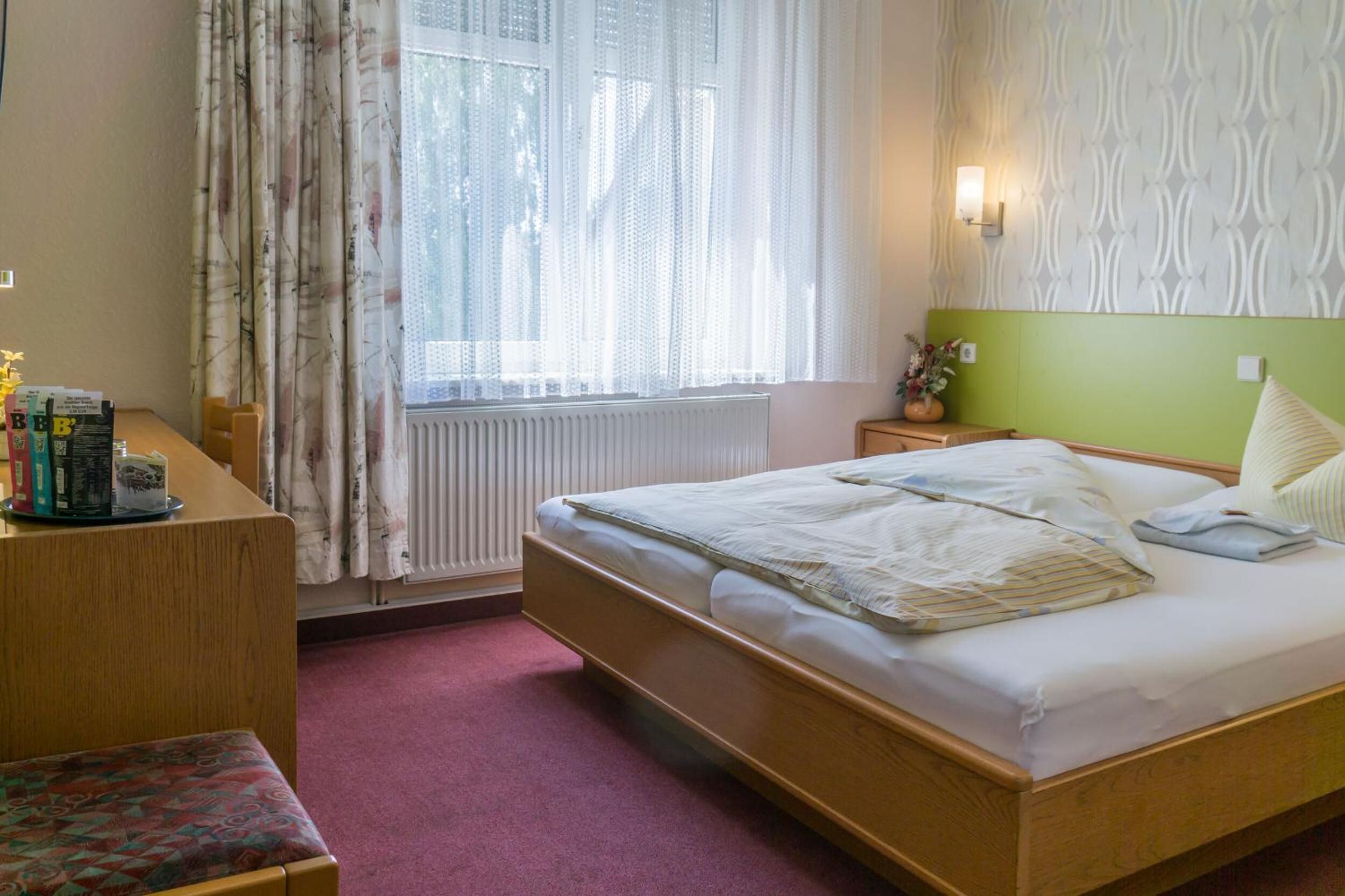 A hotel room with a wooden bed, a desk with books, a window with sheer curtains, and a patterned wall.