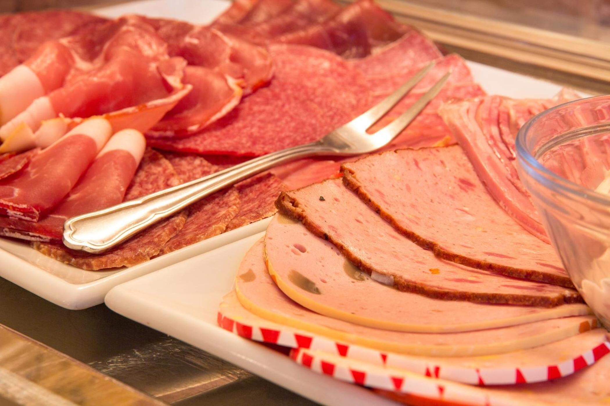 Una variedad de embutidos en un plato, incluyendo jamón y salami, con un tenedor y un bol de vidrio cerca.
