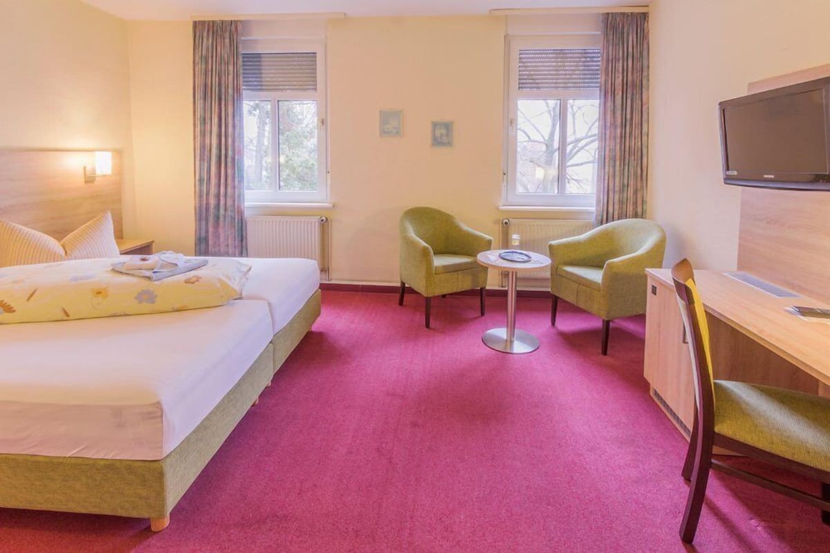 A hotel room with a bed, two armchairs, a small table, a desk, and a TV.