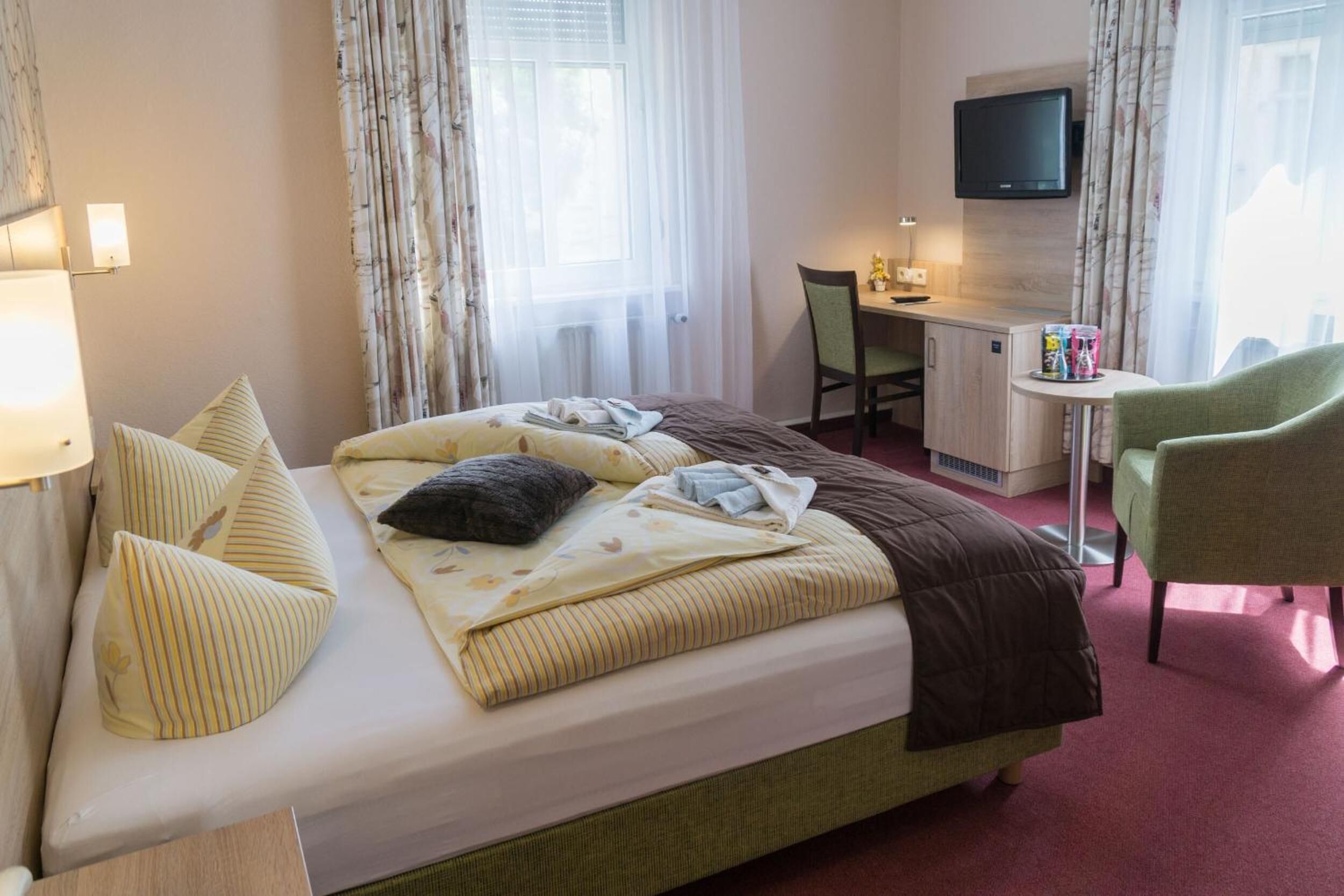 A hotel room with a bed, a desk, a chair, a TV, and a small table with a lamp.