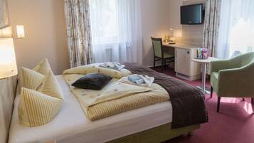 A hotel room with a bed, a desk, a chair, a TV, and a small table with a lamp.