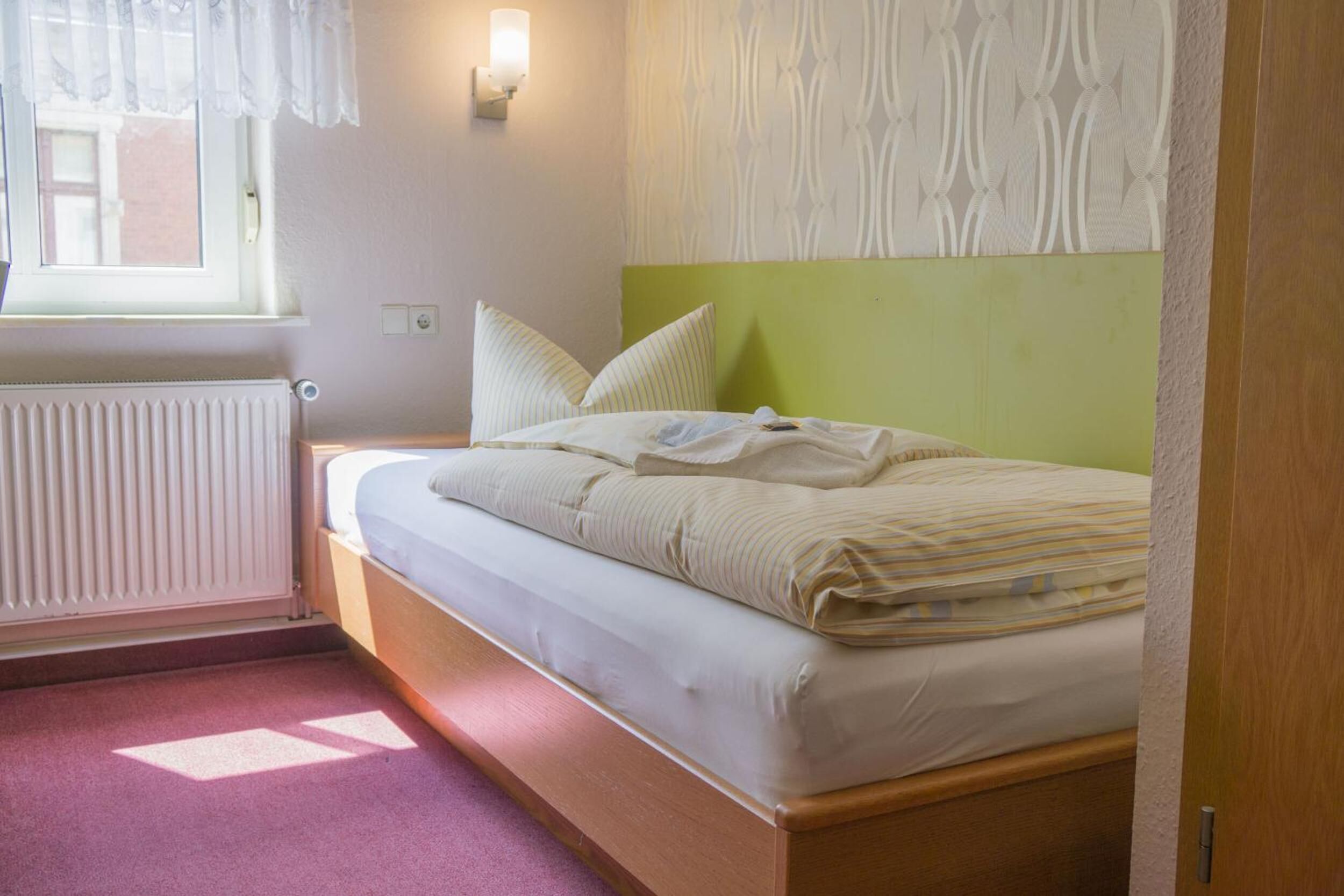 Una cama individual con cabecera verde, una ventana con cortina blanca y una lámpara fijada a la pared.