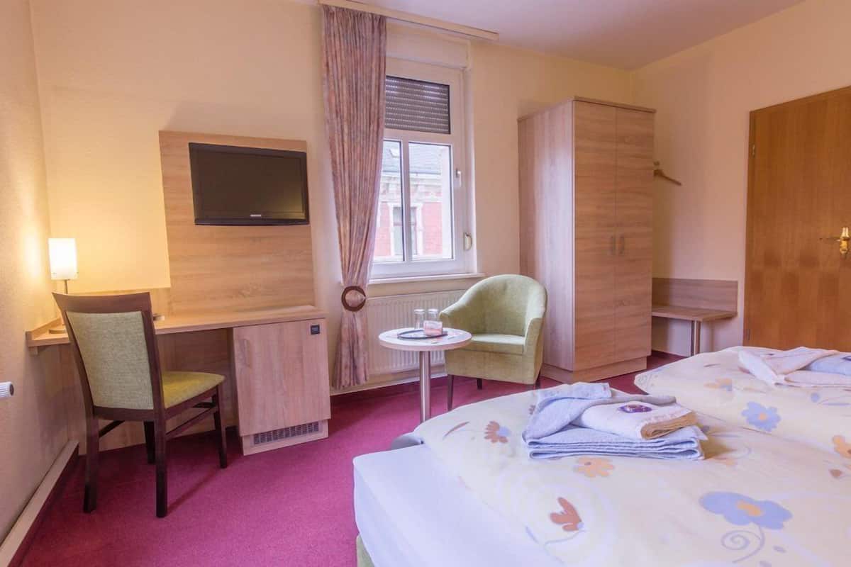 A hotel room with a bed, a desk with a chair, a television, a wardrobe, and a window with curtains.