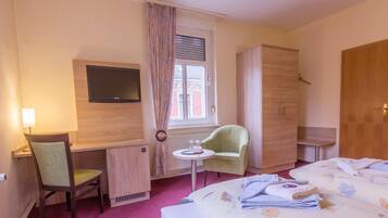 Une chambre d’hôtel comprenant un lit, un bureau avec une chaise, une télévision, une armoire et une fenêtre avec des rideaux.