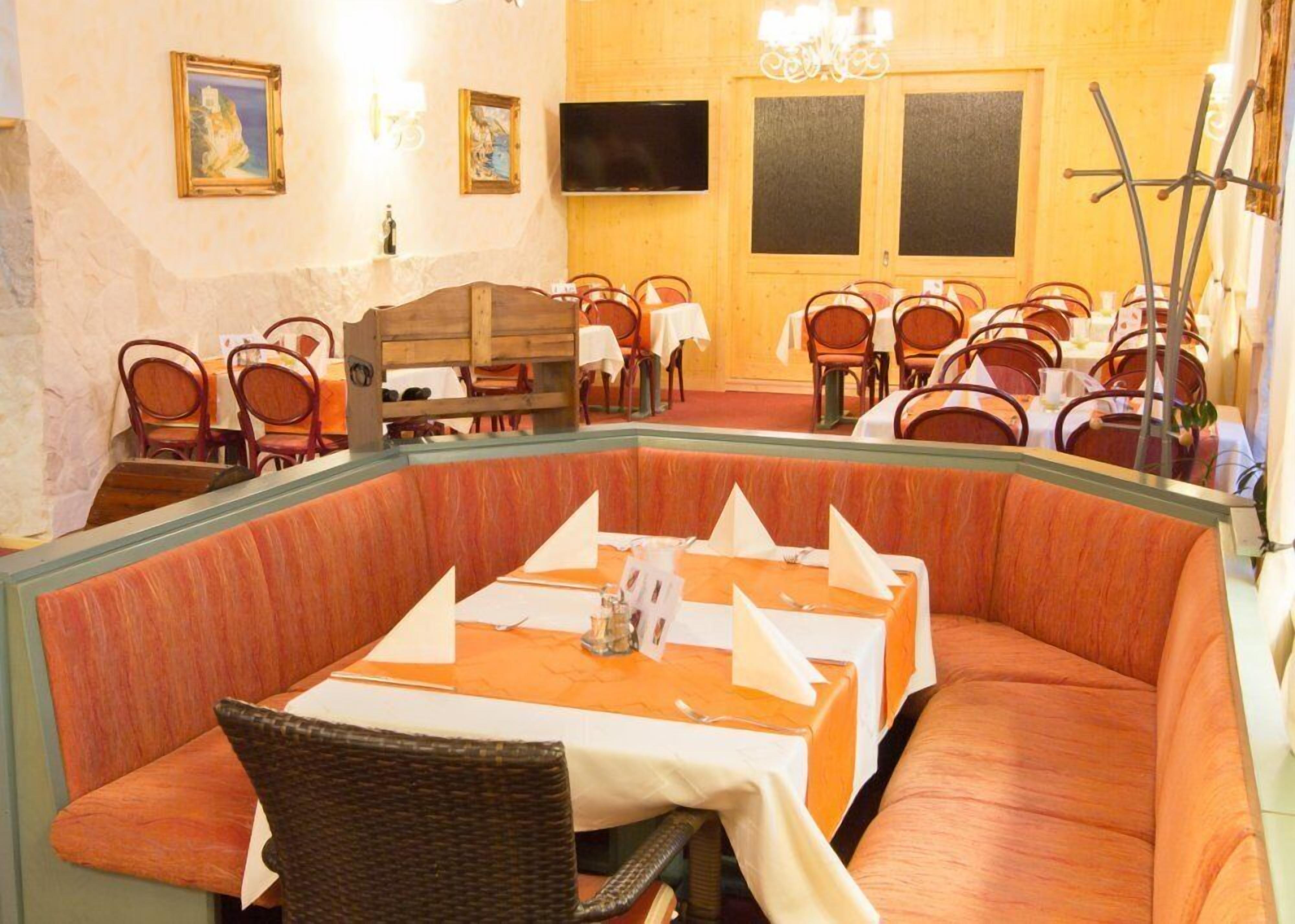 A restaurant with a central seating area featuring a long table with white napkins and a bottle of water. There are multiple tables and chairs arranged around the room, and a television mounted on the wall.