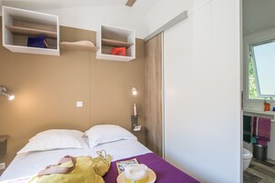 Una habitación de hotel moderna con una cama, dos almohadas blancas, una manta morada, un bolso amarillo y un sombrero sobre la cama. Hay dos estantes blancos sobre la cama, uno con un bolso azul y otro con un bolso rojo. Una botella blanca está sobre un estante pequeño al lado de la cama.