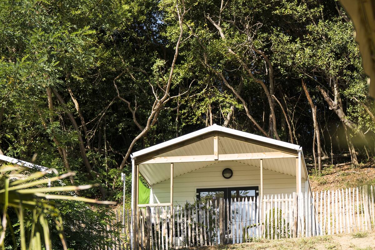 Ein kleines, eingeschossiges Gebäude mit überdachter Veranda, umgeben von einem Holzzaun und dichter Baumvegetation.