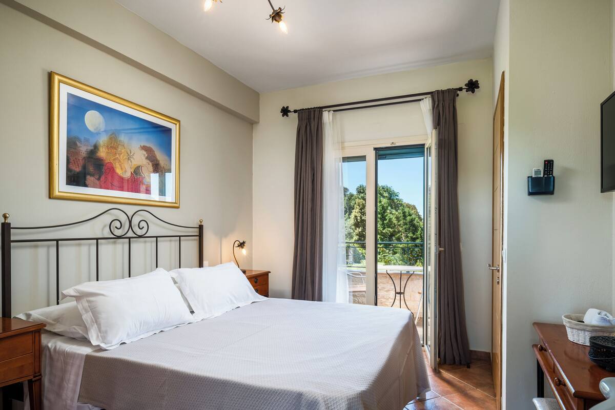 Ein Schlafzimmer mit Bett, Nachttisch, Kommode, einem Gemälde, einem Fenster mit Vorhängen sowie einem Balkon mit Tisch und Stühlen.