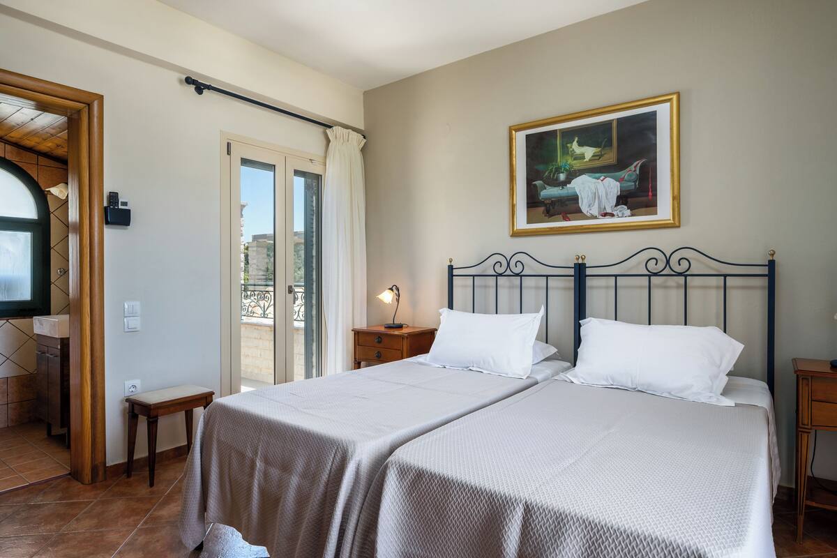 Ein Schlafzimmer mit Bett, Nachttisch, einem Fenster mit Vorhängen, einem Bild an der Wand und einer Tür zu einem anderen Raum.