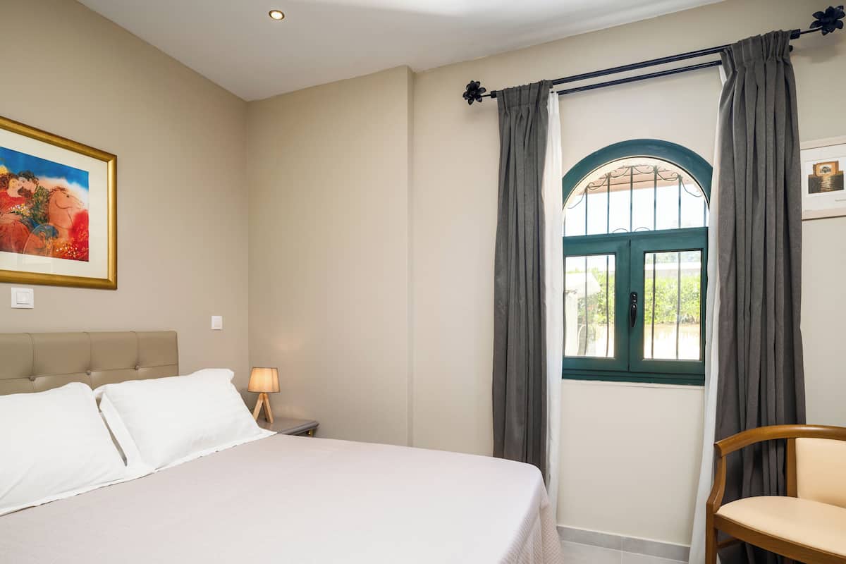 Ein Schlafzimmer mit einem Bett, einem Nachttisch, einem Sessel, einem Fenster mit Vorhängen und einem Bild an der Wand.