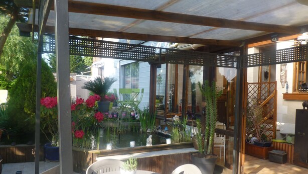 Een overdekt terras met een ronde tafel en stoelen, omringd door potplanten en een kleine waterpartij.