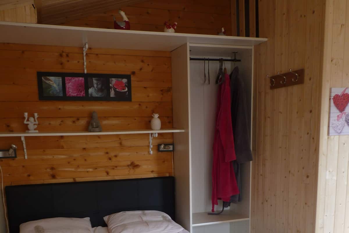 A bed with a white duvet and a black headboard, a wooden wall with a shelf above the bed, and a coat rack with a red jacket hanging on it.