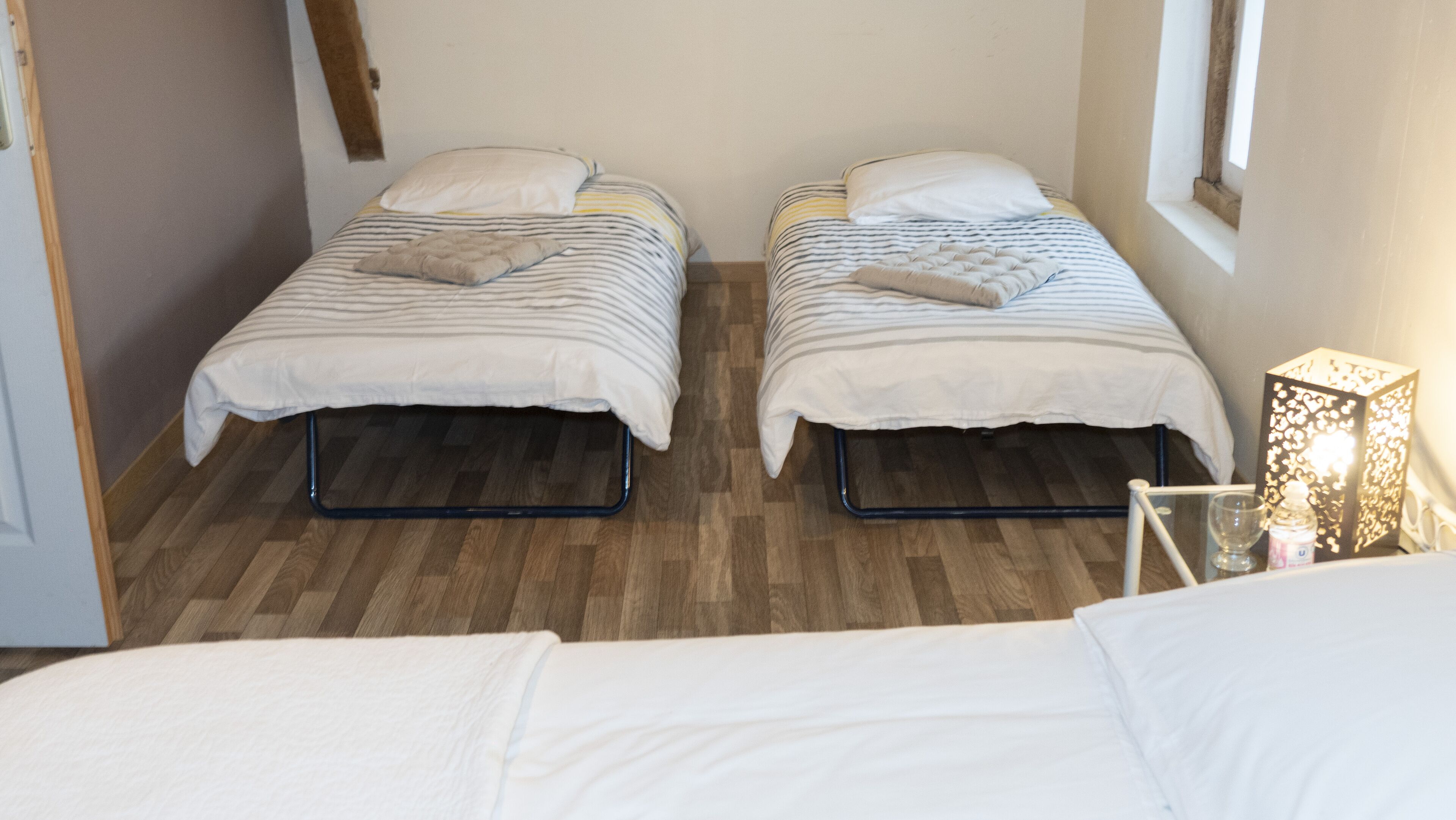 Une chambre avec deux lits simples, chacun étant équipé d’un oreiller et d’une couette. Le sol est en bois et il y a une fenêtre sur le côté droit.
