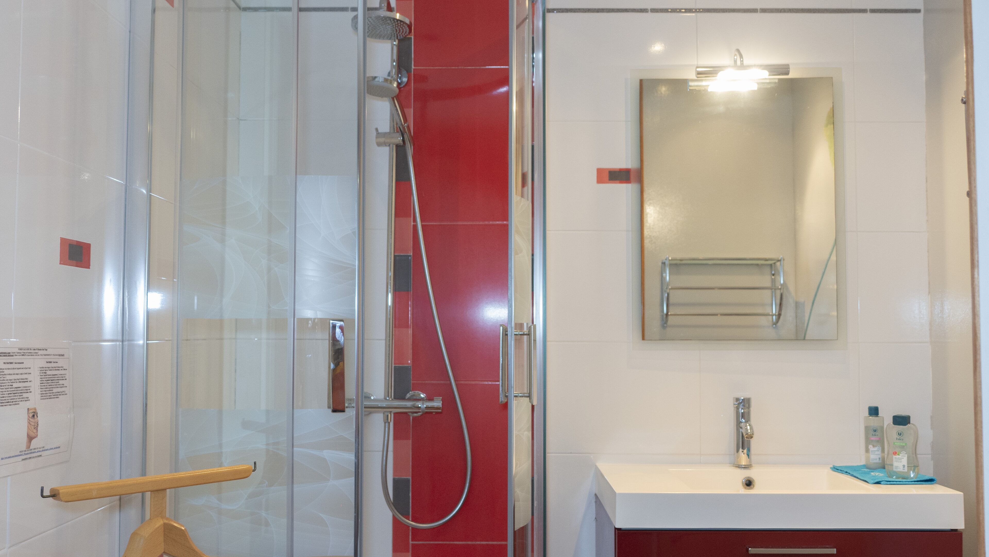A modern bathroom with a glass shower enclosure, a white sink with a mirror above it, and a red tiled wall.