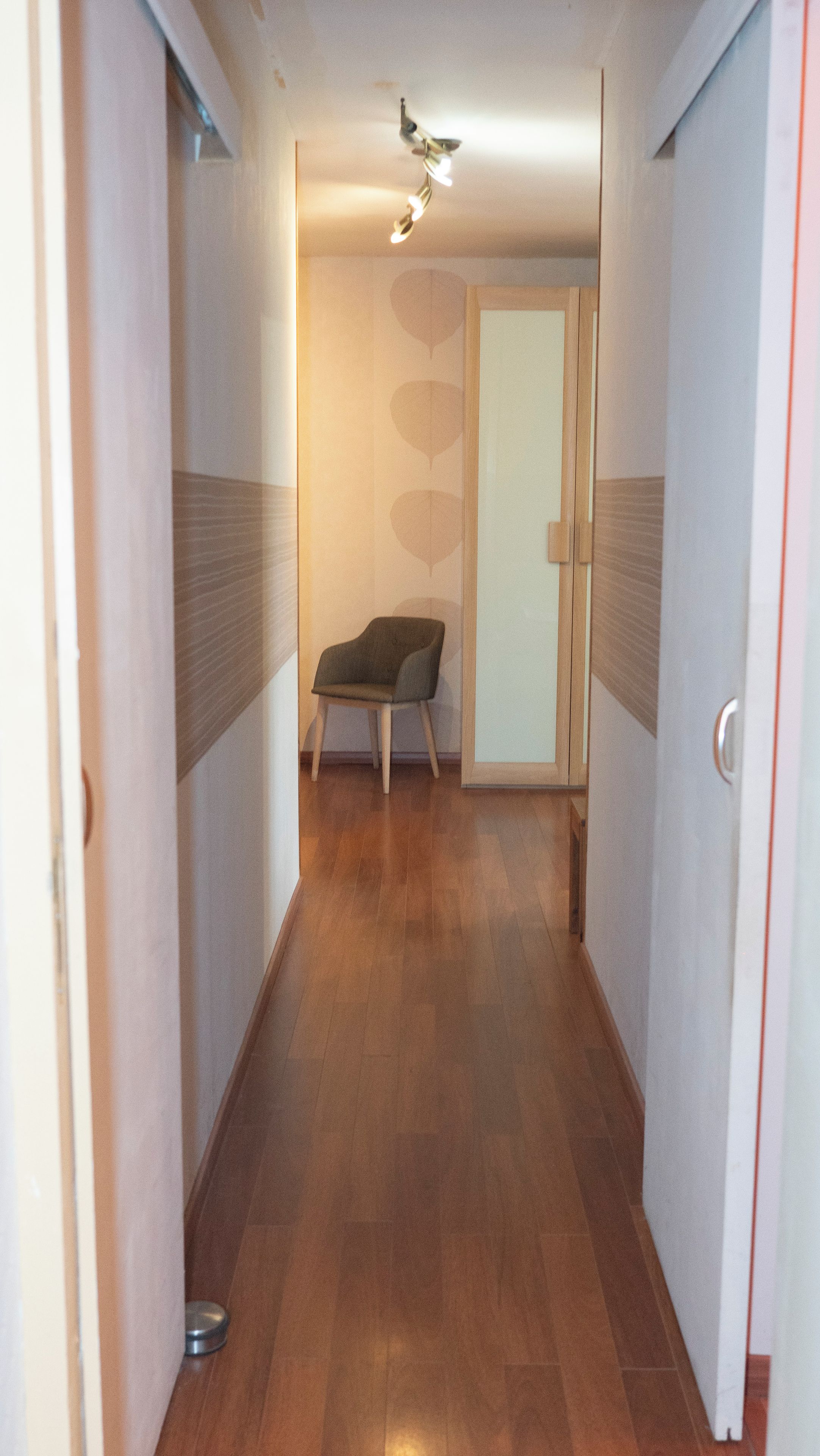 Un couloir étroit au plancher en bois, avec une chaise et une porte dotée d’un panneau en verre dépoli.