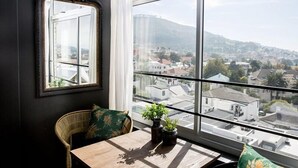 A room with a large window, a mirror, a wicker chair, a small table, and two potted plants. - On Kloof Self Catering Apartment (Cape Town)