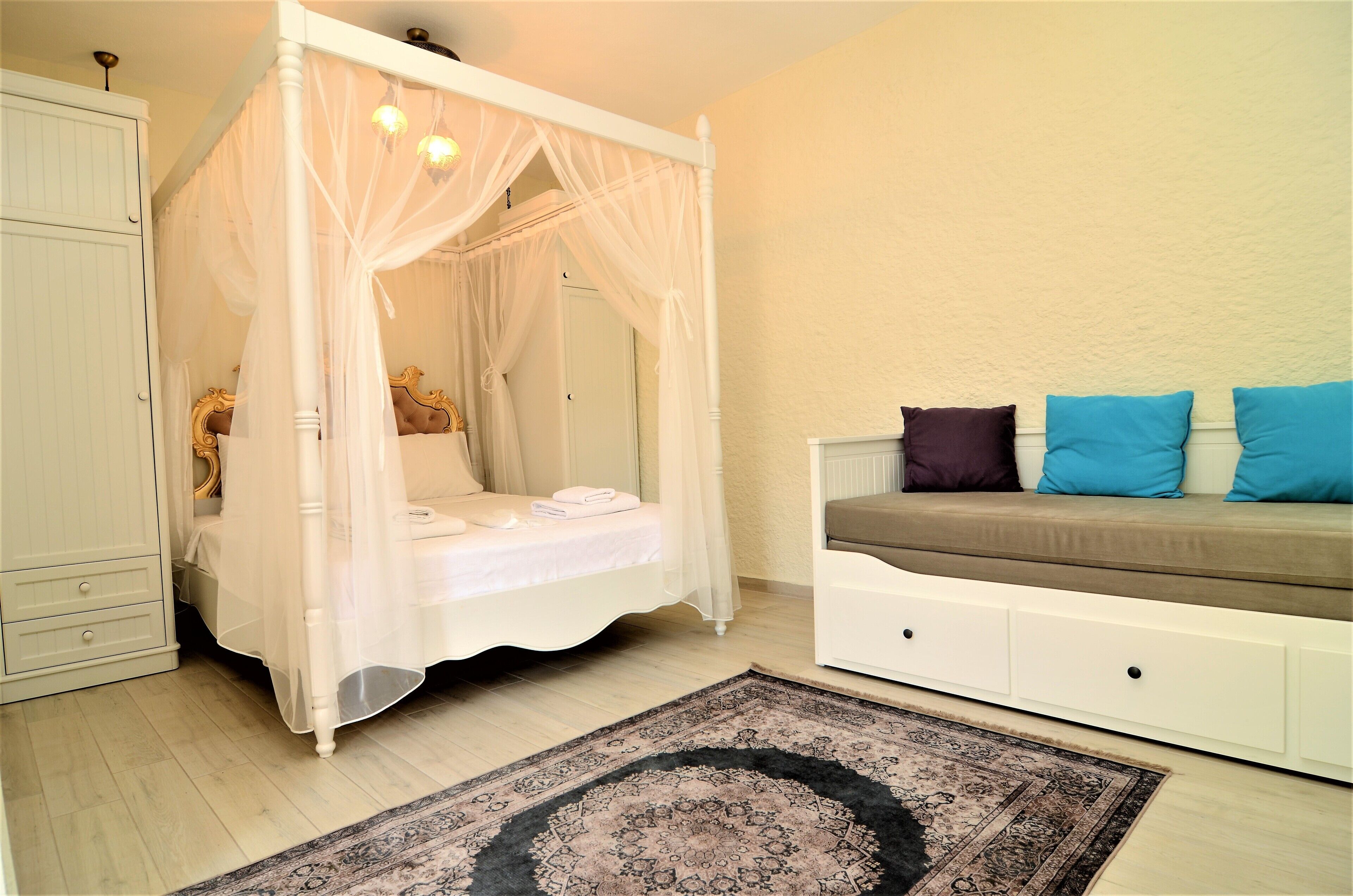 A bedroom with a four-poster bed, a sofa with cushions, a bedside table, and a wardrobe.
