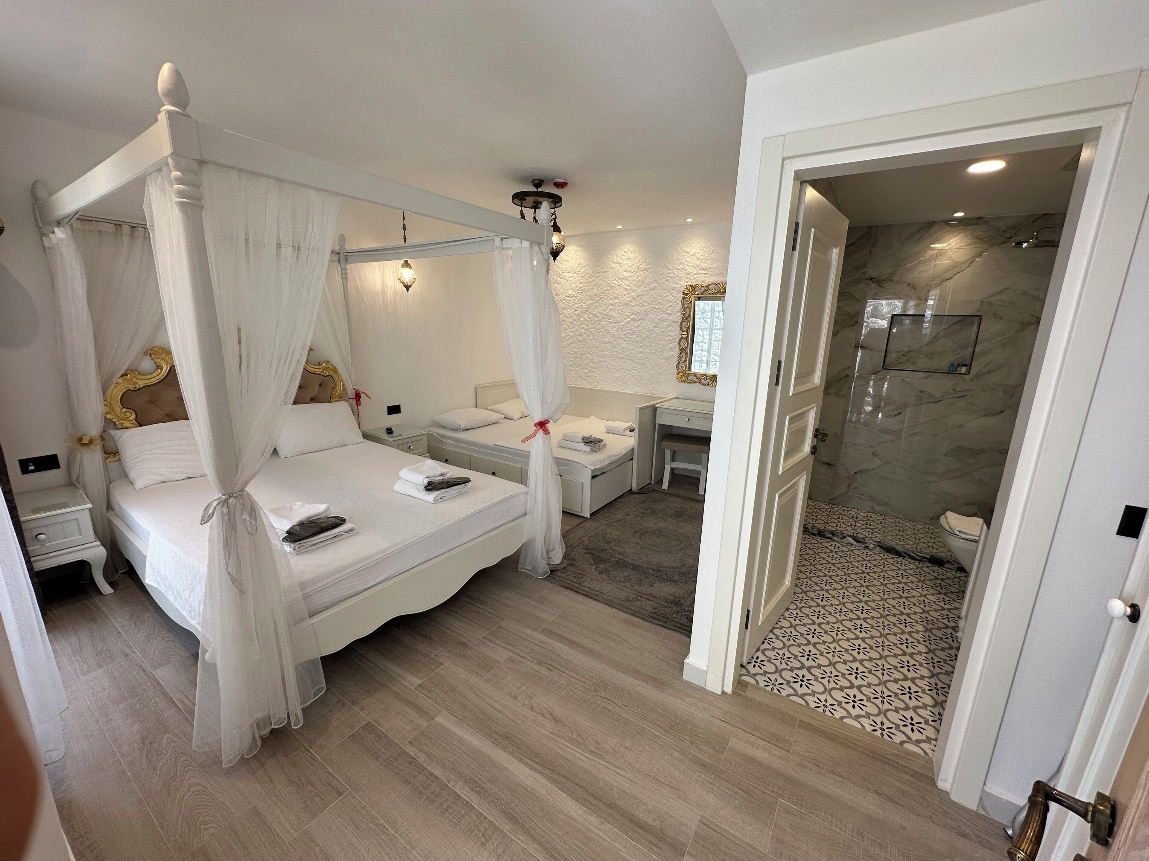 Un lit à baldaquin avec une literie blanche, une table de chevet, un petit bureau avec une chaise et un espace douche avec un mur en marbre et des carreaux de sol à motifs.