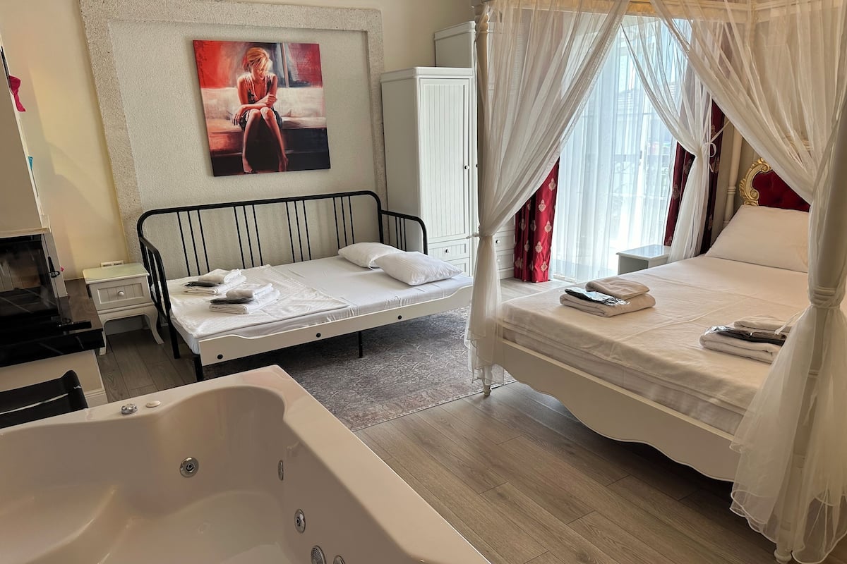 Ein Schlafzimmer mit einem Bett, einem kleinen Nachttisch, einem großen Fenster mit leichtem Vorhang, einem an der Wand befestigten Fernseher und einem Gemälde.