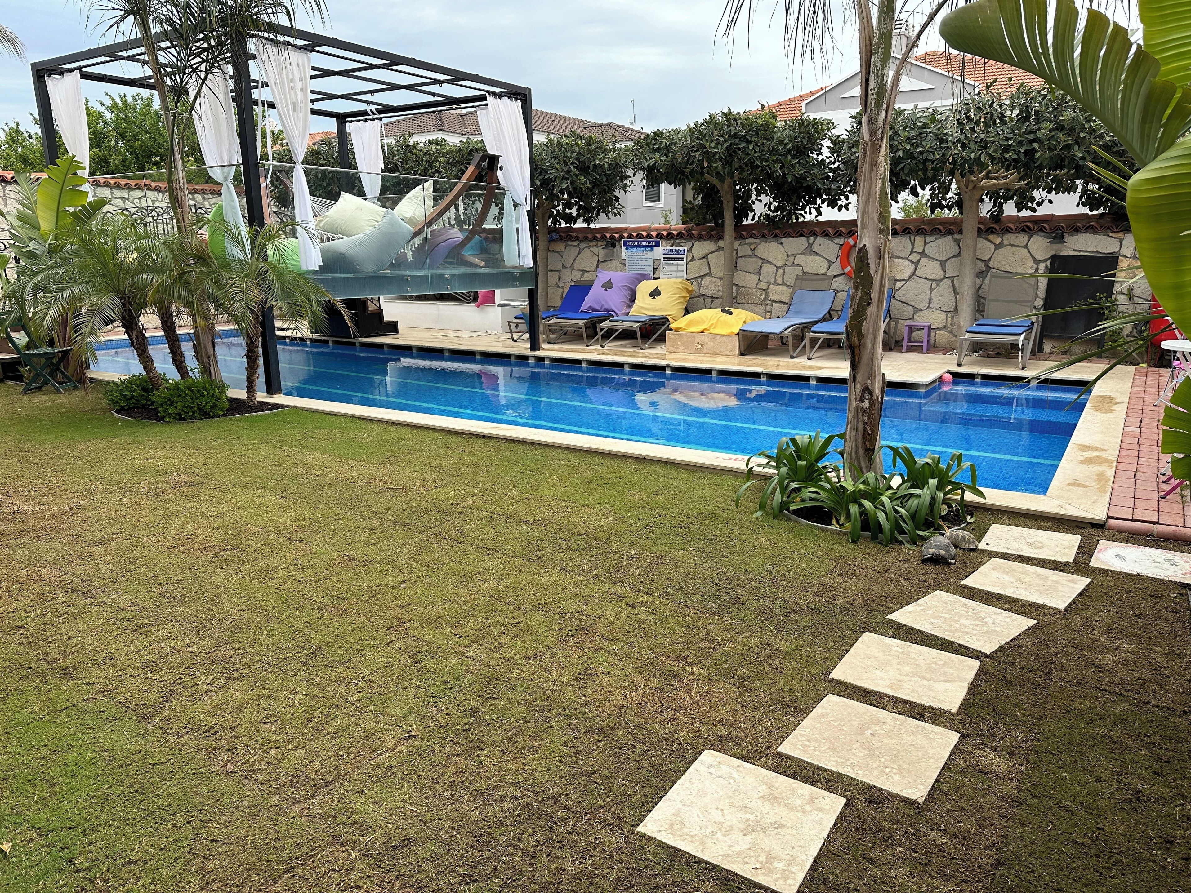 Uma piscina com espreguiçadeiras, um pergolado com cortinas brancas e um muro de pedra ao fundo.