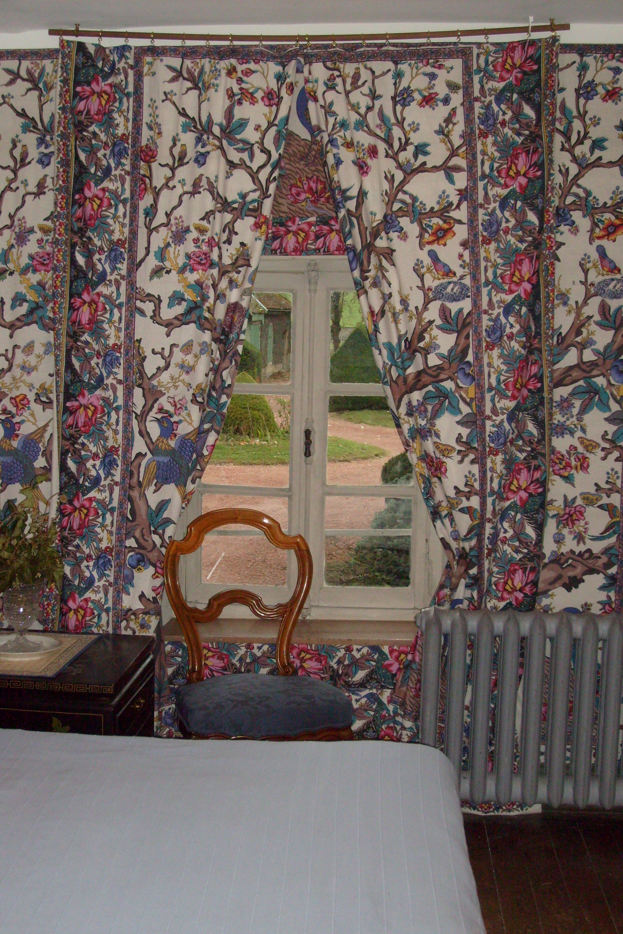 Een slaapkamer met een bed, een stoel, een radiator en een raam met bloemen gordijnen.
