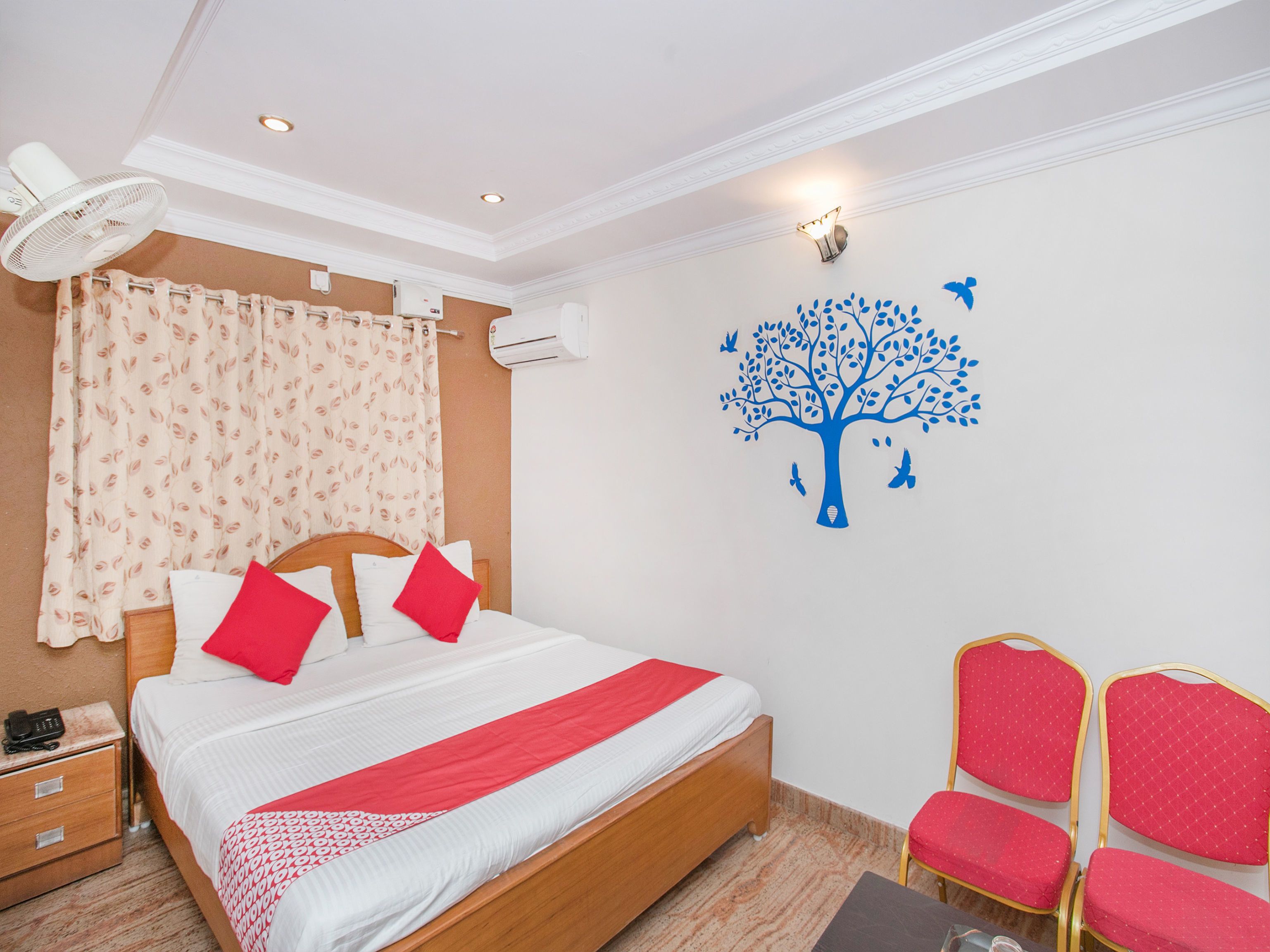 A hotel room with a bed, two red chairs, a wall-mounted tree decal, and an air conditioning unit.