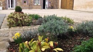 A stone building with a large garage door, a gravel driveway, and a landscaped garden with various plants. - La Ferme du Bois Paris (Ermenonville-la-Petite)