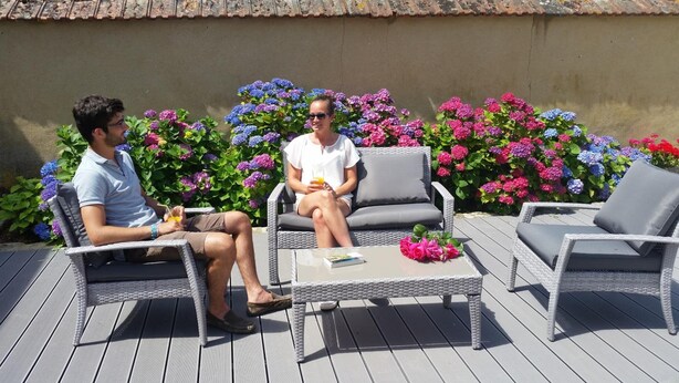 Two people seated on wicker chairs on an outdoor deck with a table in between, surrounded by vibrant flowers. - La Ferme du Bois Paris (Ermenonville-la-Petite)