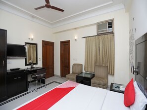 A hotel room with a bed, a desk with a TV, a chair, a small table, and a ceiling fan. - OYO 12075 Hotel Regalia Retreat (New Delhi)