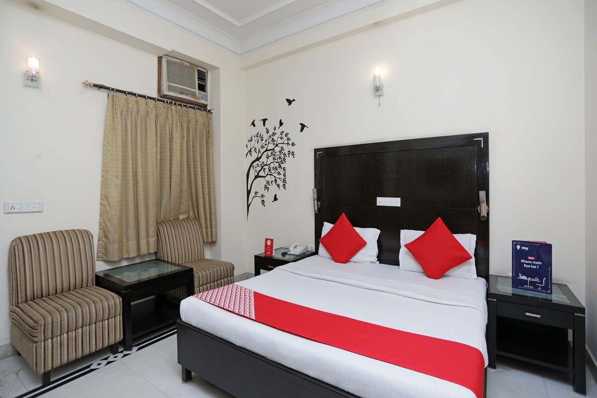 A hotel room with a bed, a chair, a nightstand, a bedside table, an air conditioning unit, and a wall decoration.